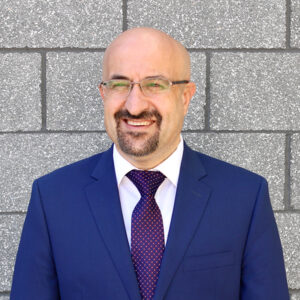 A man in a blue suit stands outside and smiles.